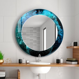 Pu&ograve; includere: Uno specchio da bagno rotondo con un design astratto blu e verde attorno al bordo. Lo specchio &egrave; montato su una parete piastrellata bianca sopra un lavandino bianco e un piano in legno.