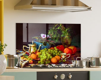 May include: A rectangular kitchen backsplash panel featuring a still life print of green grapes, red apples, and oranges arranged on a patterned cloth. A silver teapot, a small silver creamer, and a potted plant with purple flowers are included.