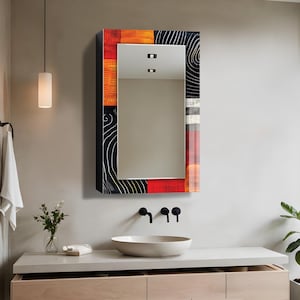 Könnte beinhalten: Ein rechteckiger Wand-Medizinschrank mit einer Spiegeltür und einem dekorativen Rahmen. Der Rahmen zeigt ein mehrfarbiges abstraktes Muster mit Abschnitten in Orange, Schwarz mit weißen Liniendetails und durchgehenden roten vertikalen Streifen.