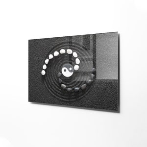 May include: A black and white yin yang symbol is placed in the center of a black sand zen garden. White stones are arranged in a spiral pattern around the symbol.