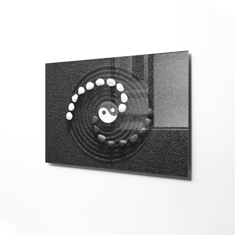 May include: A black and white yin yang symbol is placed in the center of a black sand zen garden. White stones are arranged in a spiral pattern around the symbol.