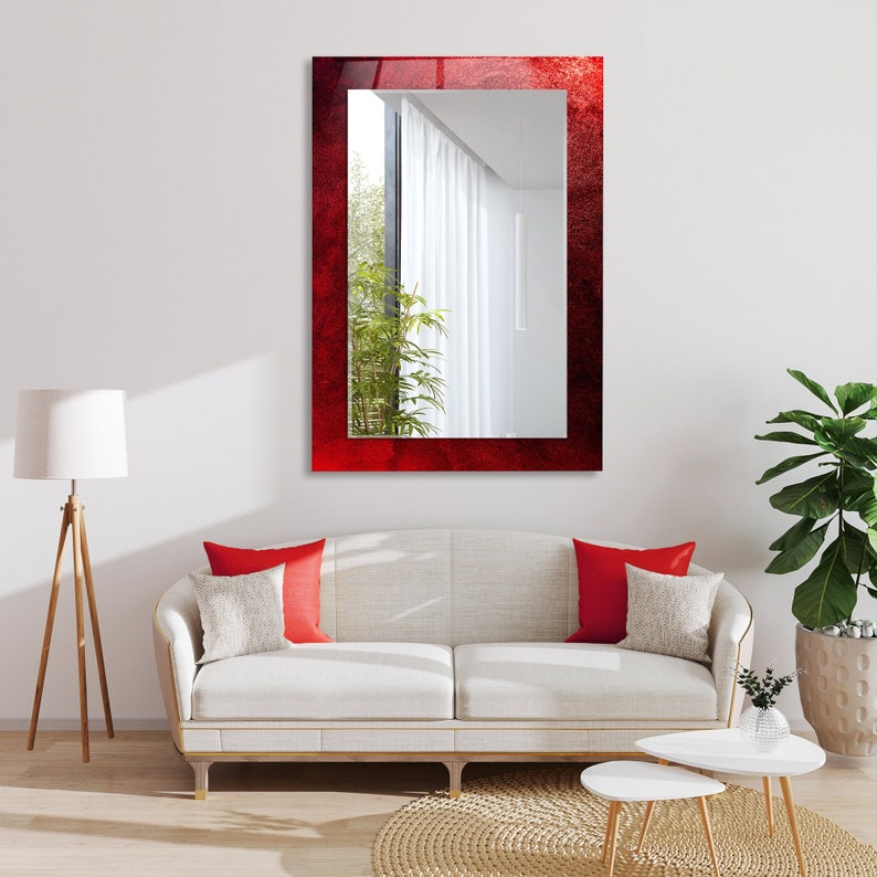 K&ouml;nnte beinhalten: Ein roter Spiegel mit einem strukturierten Rahmen h&auml;ngt an einer wei&szlig;en Wand &uuml;ber einem wei&szlig;en Sofa mit roten Kissen. Der Spiegel spiegelt einen Raum mit einem Fenster und wei&szlig;en Vorh&auml;ngen wider.