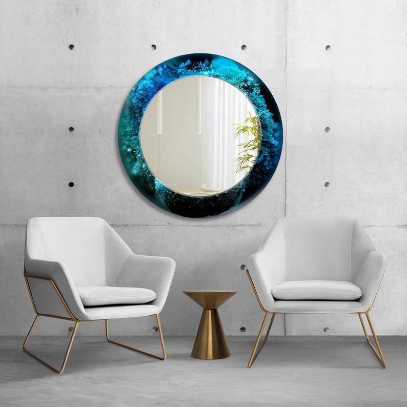 Pu&ograve; includere: Uno specchio rotondo con un design astratto blu e verde. Lo specchio &egrave; appeso a un muro di cemento grigio. Due sedie bianche con telai dorati sono davanti allo specchio. Un piccolo tavolino da salotto dorato si trova tra le sedie.