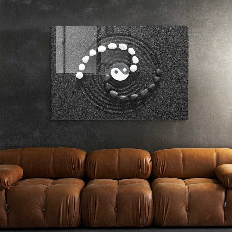 May include: A black and white yin yang symbol is surrounded by a spiral of white and black stones on a black sand background. The image is framed and hanging on a gray wall above a brown leather couch.