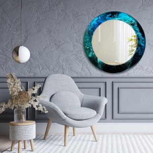 Pu&ograve; includere: Uno specchio rotondo con una cornice in resina blu e verde. Lo specchio &egrave; appeso a una parete grigia sopra una poltrona grigia.