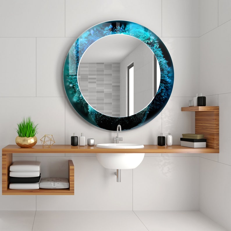 Pu&ograve; includere: Uno specchio da bagno rotondo con un design astratto blu e verde attorno al bordo. Lo specchio &egrave; montato su una parete piastrellata bianca sopra un lavandino bianco e un piano in legno.
