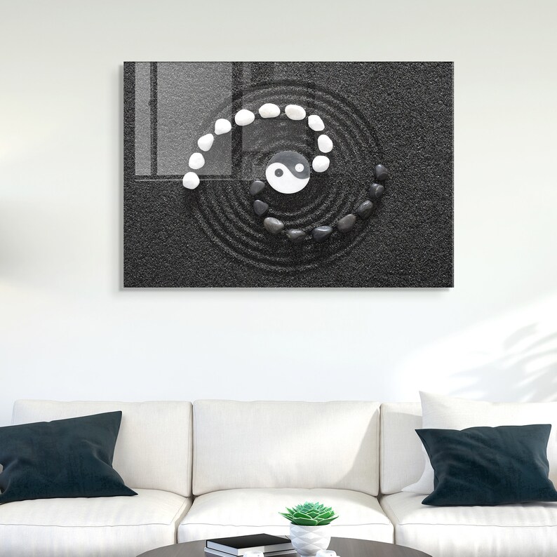 May include: A black and white yin yang symbol is surrounded by a circle of white and black stones in a zen garden design. The stones are arranged in a spiral pattern on black sand.