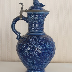 May include: A blue ceramic pitcher with a pewter lid and a detailed design of leaves and vines. The pitcher has a handle and a spout.