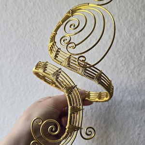 Peut inclure: Un bracelet de bras en fil métallique doré présentant un motif en spirale. La pièce est composée de plusieurs brins de fil métallique enroulés et torsadés en deux sections circulaires distinctes reliées par une structure centrale ornée.