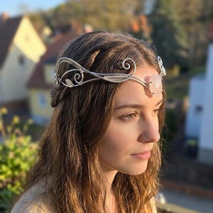Könnte beinhalten: Ein silberfarbener Draht-Haarreif mit einem zentralen Rosenquarzstein. Das Kopfstück hat zarte Wirbel und blattartige Details. Geeignet für besondere Anlässe oder Fantasy-Veranstaltungen.