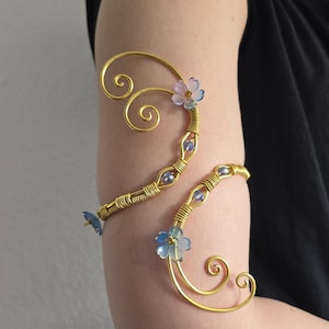 May include: A gold-toned wire arm cuff featuring swirling metal designs, small blue and purple faceted beads, and translucent light blue flower-shaped accents. The piece wraps around the upper arm with a flexible, open-ended metal structure.