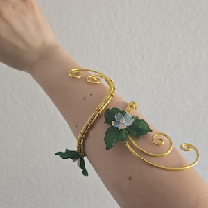 May include: A gold-toned wire arm cuff featuring a spiraling design with wrapped wire detailing. The piece is adorned with small, translucent light blue flower blossoms and deep green leaves attached to the metal frame at two distinct points.