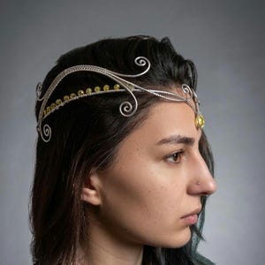 May include: A silver-toned wirework headpiece with a delicate design. The headpiece features swirling accents and small yellow gemstones. A larger yellow gemstone hangs from the center. The headpiece is worn on dark hair.