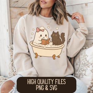 Op de afbeelding: Beige sweatshirt met een cartoon spook in een badkuip die een boek leest, met een zwarte kat in de buurt. Het spook draagt een roze strik. De tekst "HIGH QUALITY FILES PNG & SVG" staat onderaan.