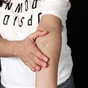 Op de afbeelding: Een kleine, minimalistische zwarte lijntekening als tijdelijke tatoeage van een pijlstaartrog op de binnenkant van de onderarm. Het ontwerp toont een eenvoudige omtrek van het lichaam en de staart in één doorlopende lijn op de huid.