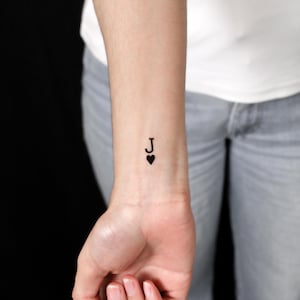 May include: A temporary tattoo on a wrist featuring a black letter J positioned directly above a small black heart symbol. The design mimics the aesthetic of a playing card suit, specifically representing a jack of hearts in a minimalist style.