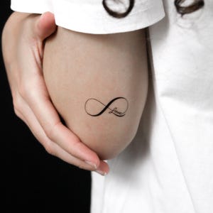 Peut inclure: Un tatouage temporaire noir représentant un symbole infini avec le mot Love écrit en écriture cursive intégré dans la boucle droite du motif. Le tatouage est appliqué sur la peau, affichant une esthétique à l'encre noire nette et épurée.