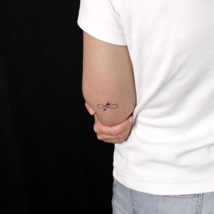 May include: A small, black ink tattoo on the skin featuring an infinity symbol intertwined with an arrow. The design is positioned on the upper arm, just above the elbow, and is rendered in a fine-line style with clean, precise black lines.