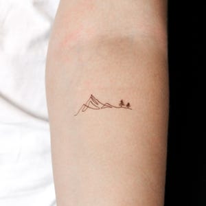 Könnte beinhalten: Ein minimalistisches Tattoo-Design mit einer Bergkette und zwei kleinen Bäumen. Die feine Linienkunst ist in schwarzer Tinte gehalten und schafft eine einfache, aber detaillierte Landschaftsszene. Das Tattoo befindet sich auf dem Arm.