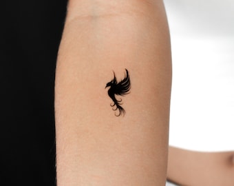 May include: A black phoenix temporary tattoo. The tattoo features a stylized bird with outstretched wings and a flowing tail. The design is a silhouette, with fine lines and details.