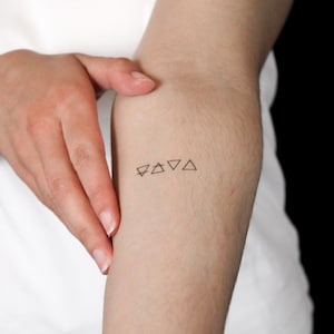 May include: A small temporary tattoo on skin featuring four distinct black line art symbols arranged in a horizontal row. The design consists of a downward-pointing triangle with a horizontal line, a triangle with a horizontal line, and two plain triangles.