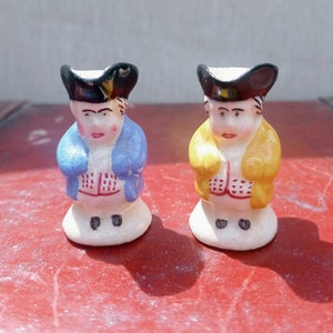 Könnte beinhalten: Zwei kleine, antike Keramikfiguren von Männern in traditioneller Kleidung. Eine Figur trägt einen blauen Mantel, die andere einen gelben Mantel. Beide haben schwarze Hüte und weiße Hemden. Die Figuren stehen auf einer roten Oberfläche.