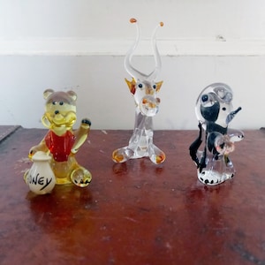 May include: Three glass figurines on a brown surface. The figurine on the left is a yellow bear holding a white bag that says "honey". The middle figurine is a clear glass animal with horns. The figurine on the right is a clear glass penguin.