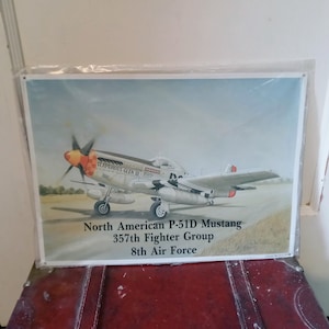 May include: A framed print of a North American P-51D Mustang fighter plane. The aircraft is silver with red and orange accents. Text on the print reads "Glamorous Glen III", "North American P-51D Mustang", "357th Fighter Group", and "8th Air Force."