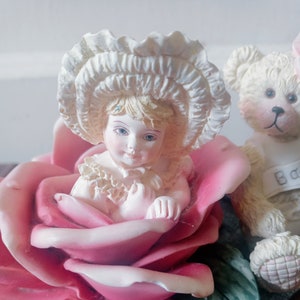 Könnte beinhalten: Eine Porzellanfigur eines kleinen Mädchens, das einen weißen Hut trägt und in einer rosa Rose sitzt. Ein Marienkäfer sitzt auf dem Rosenblatt. Ein Teddybär mit dem Wort "Baby" auf seiner Brust sitzt hinter der Rose.
