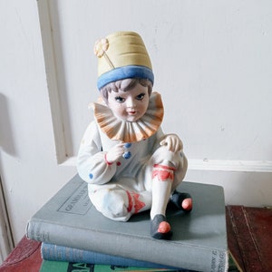 May include: A porcelain figurine of a clown with a yellow hat, blue and white ruffled collar, and red and white polka dot pants. The clown is sitting on a stack of books.