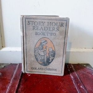May include: A vintage "Story Hour Readers Book Two" book with a beige cover. The cover features an illustration of a woman and a bear. The book is on a red surface.