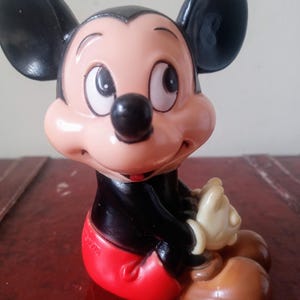May include: A vintage plastic Mickey Mouse figurine. The figurine is sitting with its legs crossed and has a red and white outfit. The figurine is brown, black, white, and red.