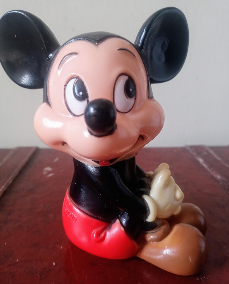 May include: A vintage plastic Mickey Mouse figurine. The figurine is sitting with its legs crossed and has a red and white outfit. The figurine is brown, black, white, and red.