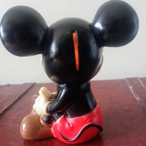 May include: A black plastic Mickey Mouse piggy bank with red shorts, brown shoes, and a slot on the back of his head.
