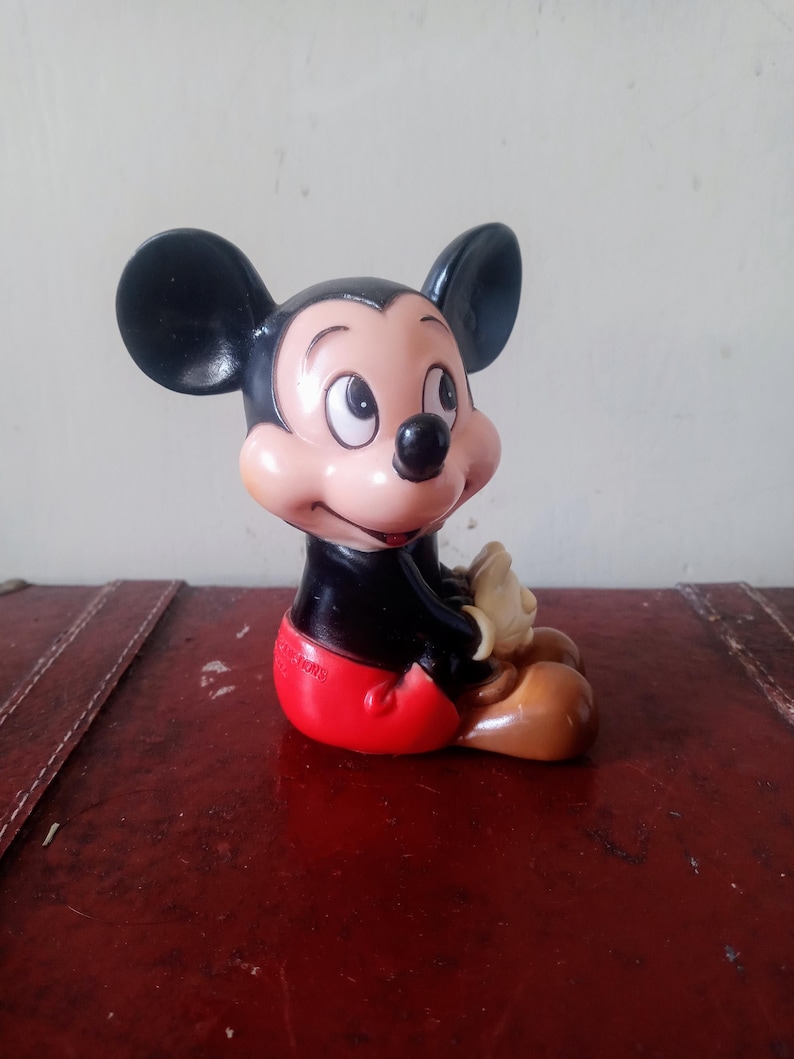 May include: Vintage Mickey Mouse figurine, seated position.  Features black ears, a light peach face, and red shorts. Brown shoes are visible. The figurine is made of plastic and shows some signs of age.  A small, partially visible marking is present on the bottom.  Classic Disney character collectible.