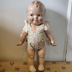 Puede incluir: Muñeca de bebé vintage con cara pintada, ojos azules y un pelele con estampado floral. La muñeca tiene un tono de piel claro y está de pie sobre una superficie estampada. Los brazos de la muñeca están ligeramente extendidos.