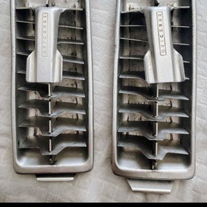 May include: Two rectangular silver-toned metal ice cube trays featuring a central lever mechanism with the word QUICKUBE embossed on the handle. Each tray has a series of horizontal metal dividers to create individual ice compartments.