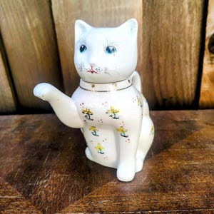 May include: A white ceramic cat-shaped teapot featuring a hand-painted pattern of small yellow flowers and red dots. The cat has blue eyes, a pink nose, and a gold-trimmed collar. One paw is extended to form the spout, and the head acts as a lid.