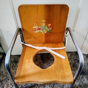 May include: A vintage wooden potty chair with a light brown finish and a cartoon illustration on the backrest. The chair has a round opening in the seat and metal legs. A white strap is attached to the seat.