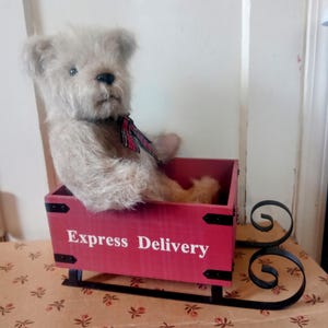 Può includere: Un orsacchiotto bianco con un fiocco rosso e nero a quadri è seduto su una slitta di legno rossa con pattini in metallo nero. La slitta ha le parole "Express Delivery" stampate sul davanti.