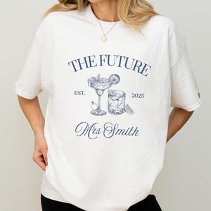 May include: Cream-coloured t-shirt featuring a navy blue graphic design. The design includes the text "THE FUTURE Mrs Smith EST. 2025" and illustrations of a margarita and a cocktail. The t-shirt has a loose fit.