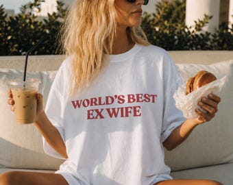 Può includere: Una t-shirt bianca a maniche corte con il testo rosso "WORLD'S BEST EX WIFE" stampato sul petto. Il capo presenta un girocollo e una vestibilità rilassata, realizzato in tessuto bianco semplice. Il testo è centrato in un carattere in grassetto.