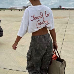 May include: A white t-shirt with the text "Found her Cowboy" in red script. The person is wearing gray cargo pants and carrying a beige tote bag. The background includes airplanes and a cloudy sky.