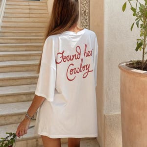 May include: White oversized t-shirt with the red text "Found her Cowboy" on the back. The shirt is worn outdoors, with a person holding a flower. The background includes stairs and a building.