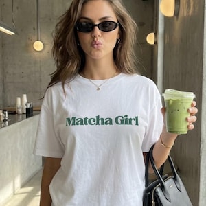 Puede incluir: Una mujer con una camiseta blanca con "Matcha Girl" en verde, pantalones de chándal grises y gafas de sol negras. Sostiene un bolso negro y una bebida verde en una taza transparente.