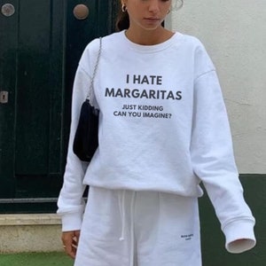 May include: White sweatshirt with the text "I hate margaritas, just kidding can you imagine?"  The model is wearing white shorts and white sneakers.