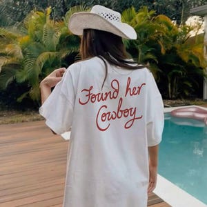 May include: White t-shirt with the red text "Found her Cowboy" on the back. A straw cowboy hat with a white band and small holes is worn. The scene is outdoors with a pool and greenery in the background.
