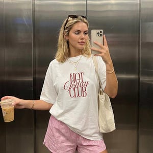May include: A white short-sleeved t-shirt with the text NOT Today CLUB printed in dark red on the chest, paired with pink and white gingham patterned shorts and a plain cream-colored canvas tote bag carried over the shoulder.