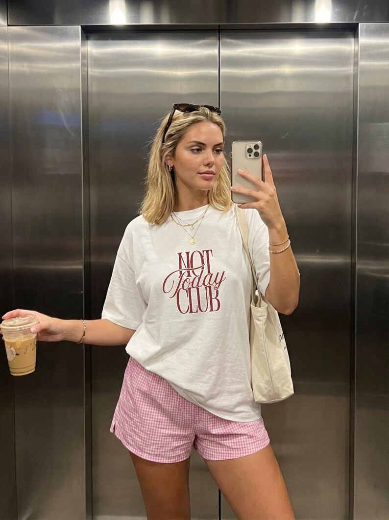 May include: A white short-sleeved t-shirt with the text NOT Today CLUB printed in dark red on the chest, paired with pink and white gingham patterned shorts and a plain cream-colored canvas tote bag carried over the shoulder.