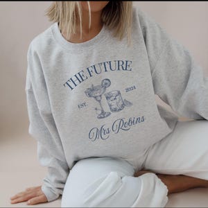 May include: A grey sweatshirt with the text "THE FUTURE EST. 2024 Mrs. Robins" and a drawing of two cocktails.
